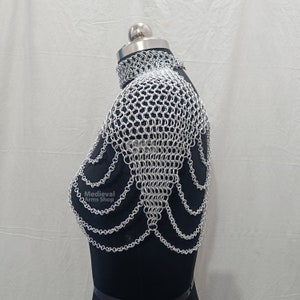 May include: Silver chainmaille shoulder piece with a decorative design. The chainmaille is made of small, interlocking metal rings. The piece is worn on a black mannequin.