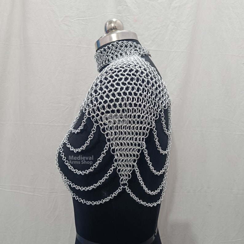 May include: Silver chainmaille shoulder piece with a decorative design. The chainmaille is made of small, interlocking metal rings. The piece is worn on a black mannequin.