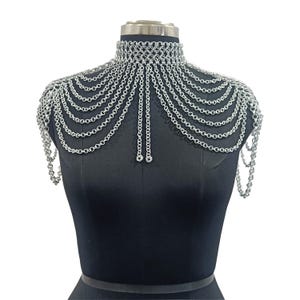 May include: A silver-toned chainmail shoulder piece featuring a high-neck choker collar constructed from interlocking metal rings. Multiple draped chain strands extend from the collar across the chest and shoulders, creating a layered effect.