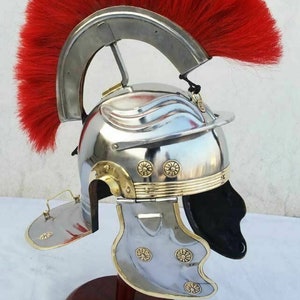 May include: A silver Roman helmet with a red horsehair crest, gold accents, and a black leather liner. The helmet is on a wooden stand.