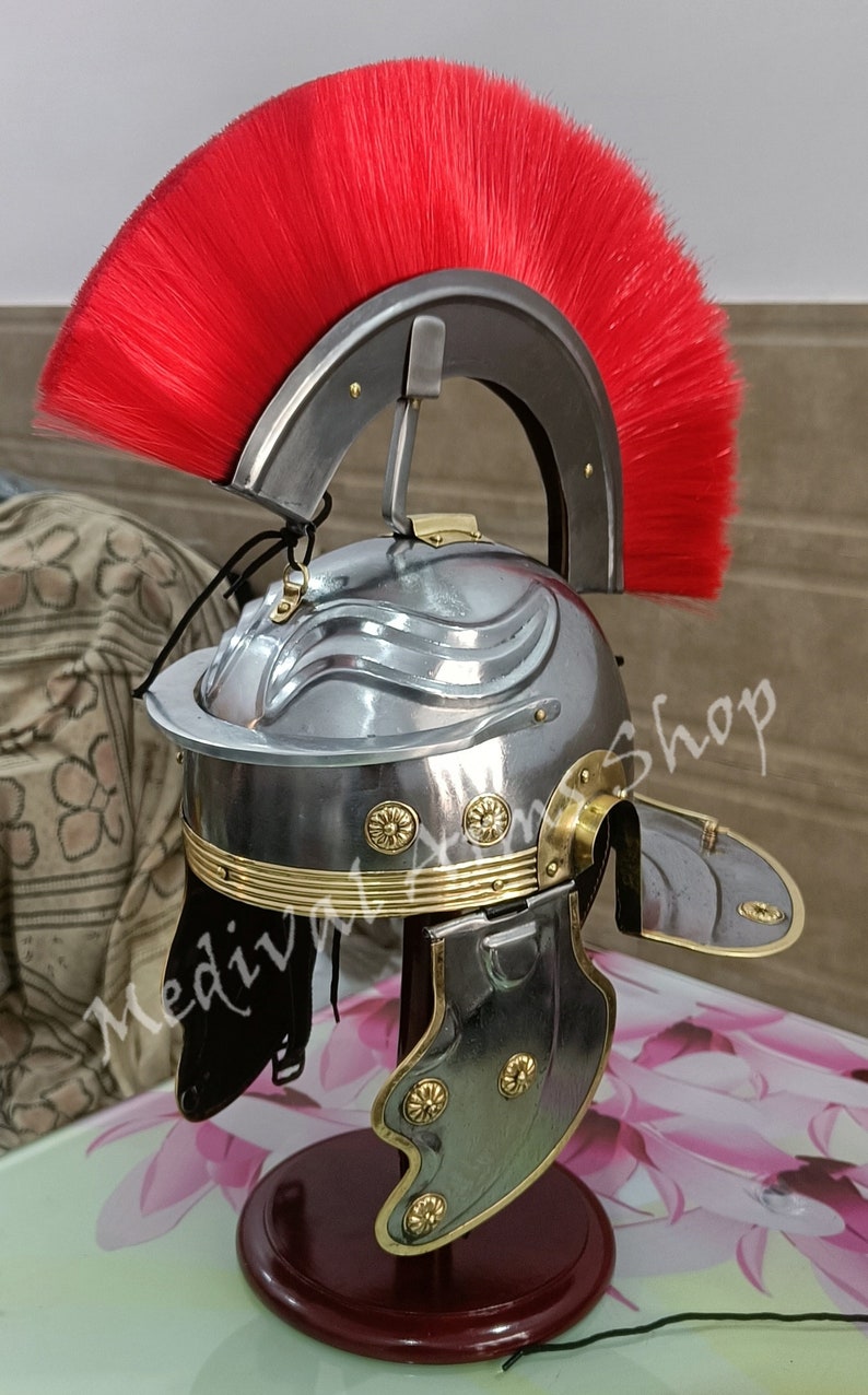 May include: A silver Roman helmet with a red horsehair crest, brass accents, and a hinged cheek guard. The helmet is displayed on a wooden stand.