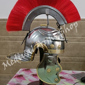 May include: A silver Roman helmet with a red horsehair crest, gold accents, and a cheek guard. The helmet is on a wooden stand.