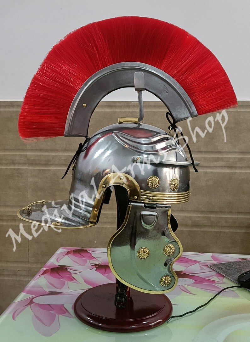 May include: A silver Roman helmet with a red horsehair crest, gold accents, and a cheek guard. The helmet is on a wooden stand.