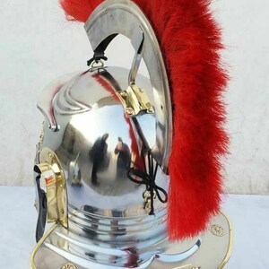 May include: A silver Roman helmet with a red horsehair crest. The helmet has a brass trim and is displayed on a wooden stand.
