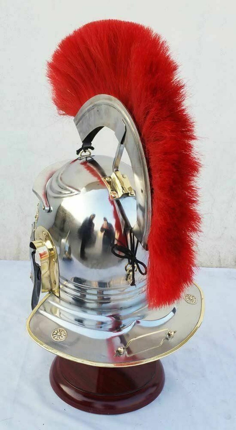May include: A silver Roman helmet with a red horsehair crest. The helmet has a brass trim and is displayed on a wooden stand.