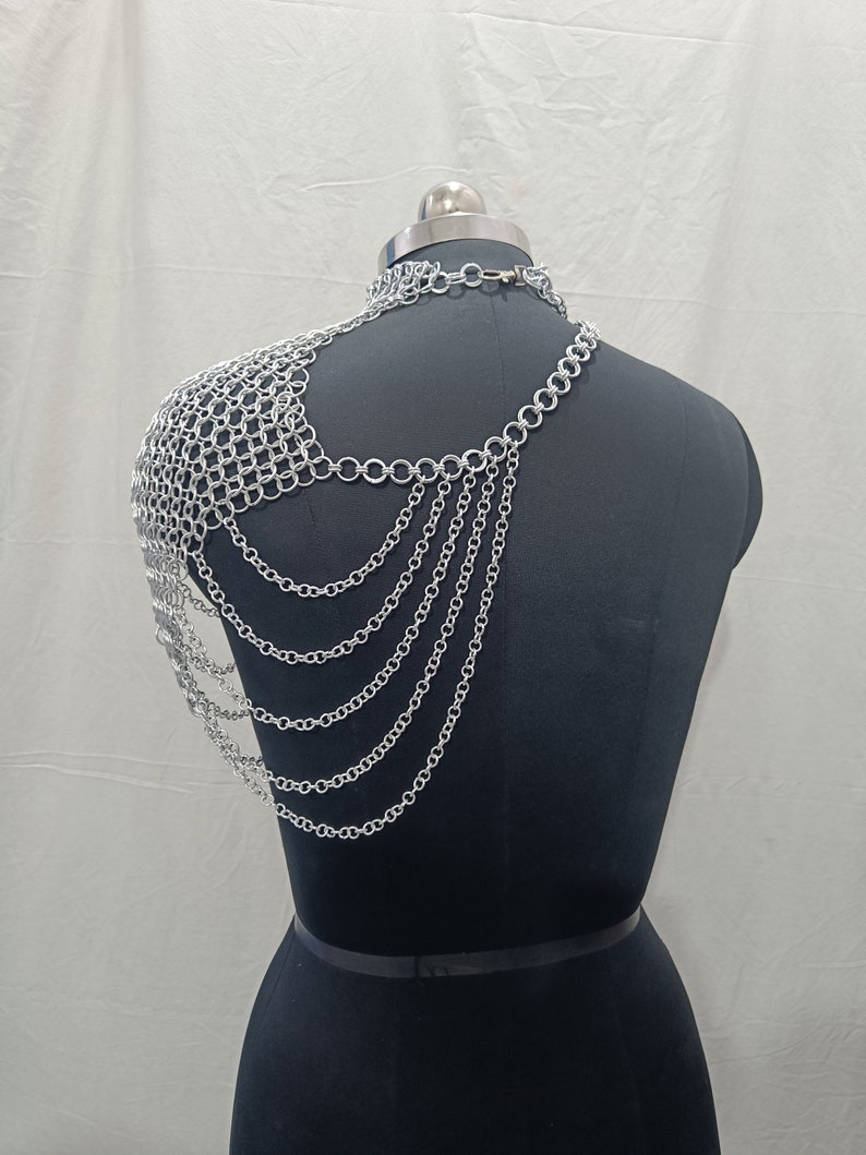 May include: Silver chainmail shoulder piece with multiple strands of chain hanging down the back. The chainmail is a delicate mesh pattern and the chains are linked together with small, round rings.