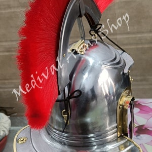 May include: A silver Roman helmet with a red horsehair crest. The helmet has a brass trim and a brass visor. The helmet is on a wooden stand.