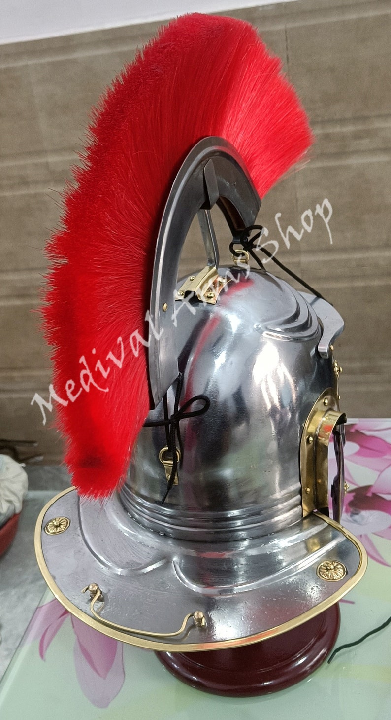 May include: A silver Roman helmet with a red horsehair crest. The helmet has a brass trim and a brass visor. The helmet is on a wooden stand.