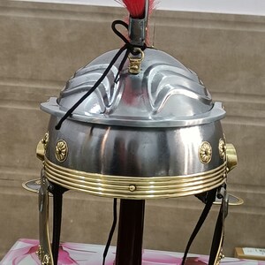 May include: A silver Roman helmet with a red plume, gold trim, and black leather straps. The helmet is on a wooden stand with a brown base.