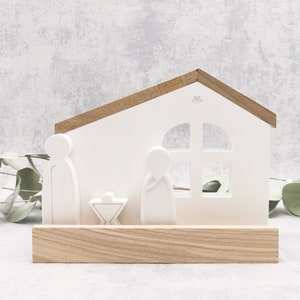 Könnte beinhalten: Weiße Holz-Krippe mit einem Stern auf dem Dach, einem Fenster und drei Figuren im Inneren. Die Szene steht auf einem Holzsockel.