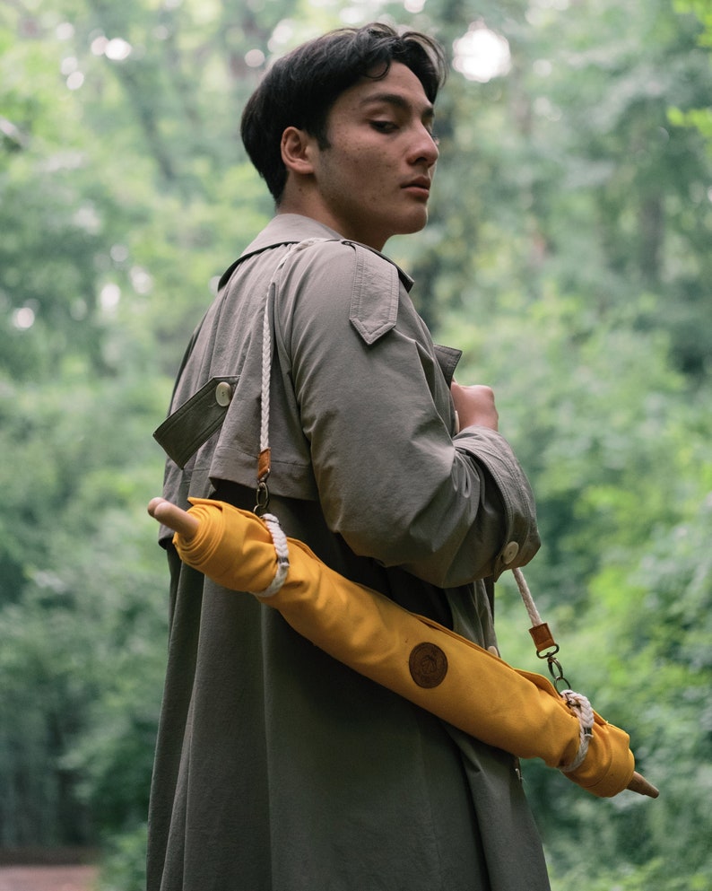 Op de afbeelding: Een persoon draagt een grijze trenchcoat met een gele canvas paraplu die over de schouder is gegespt. De paraplu heeft een bruine leren band en een witte touwband.