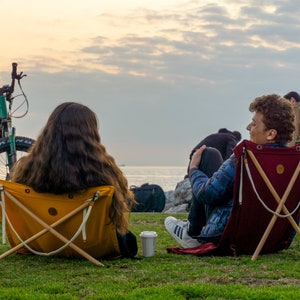 Op de afbeelding: Twee mensen zitten op opvouwbare stoelen met uitzicht op de zee. De persoon links zit op een gele stoel en de persoon rechts op een rode stoel. Een groene fiets staat op de achtergrond.