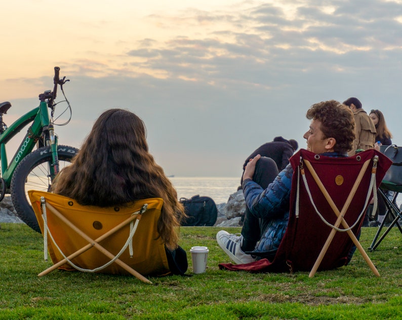 Op de afbeelding: Twee mensen zitten op opvouwbare stoelen met uitzicht op de zee. De persoon links zit op een gele stoel en de persoon rechts op een rode stoel. Een groene fiets staat op de achtergrond.