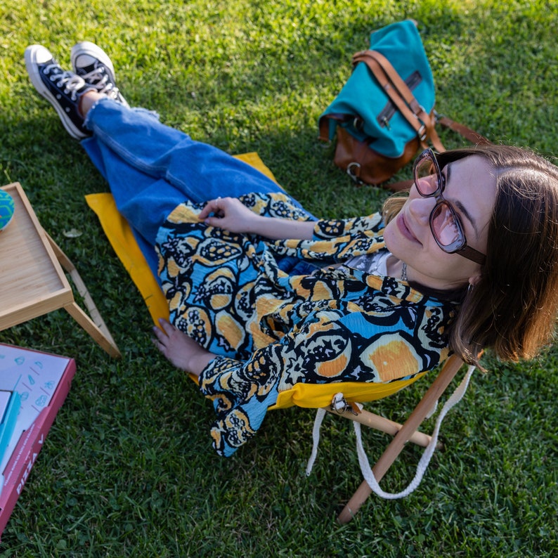 Op de afbeelding: Een vrouw in een blauwe en gele kimono zit op een gele vouwstoel met een blauwe en bruine rugzak op het gras. Ze draagt een bril en heeft haar haar naar achteren getrokken.