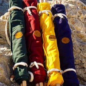 Op de afbeelding: Vier opgerolde canvas tassen met houten handvatten. De tassen zijn groen, rood, geel en blauw. Elke tas heeft een leren patch met een logo.