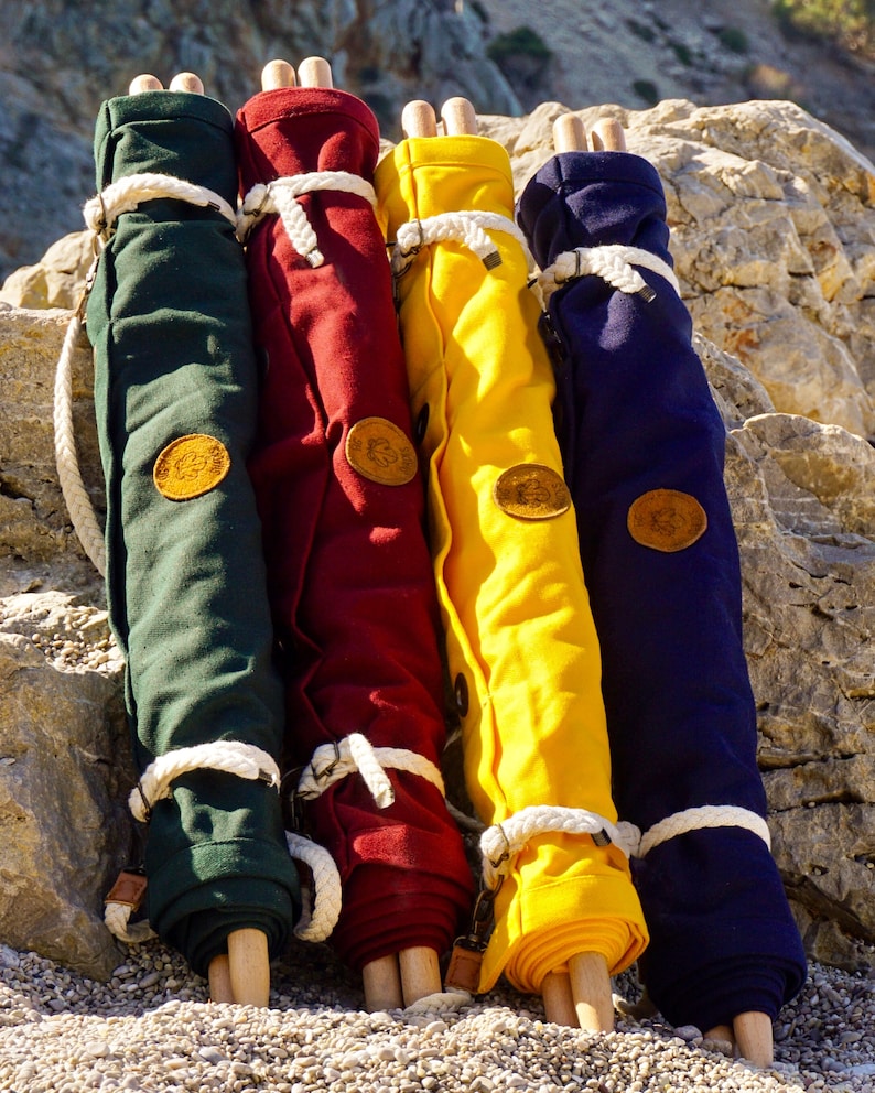 Op de afbeelding: Vier opgerolde canvas tassen met houten handvatten. De tassen zijn groen, rood, geel en blauw. Elke tas heeft een leren patch met een logo.
