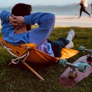 Op de afbeelding: Een persoon ontspant in een gele opvouwbare stoel met een blauwe trui en jeans. Een houten skateboard met turquoise wielen ligt op het gras.