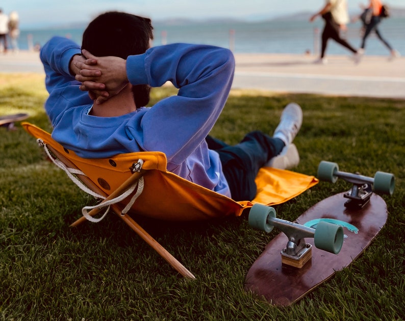 Op de afbeelding: Een persoon ontspant in een gele opvouwbare stoel met een blauwe trui en jeans. Een houten skateboard met turquoise wielen ligt op het gras.