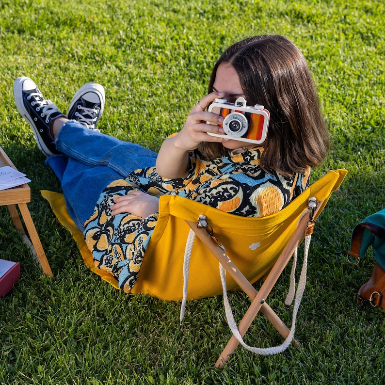 Op de afbeelding: Een persoon die op een gele vouwstoel zit en een vintage camera met een wit en rood ontwerp vasthoudt. De persoon draagt ​​een blauwe spijkerbroek en een kleurrijk, geprint shirt.