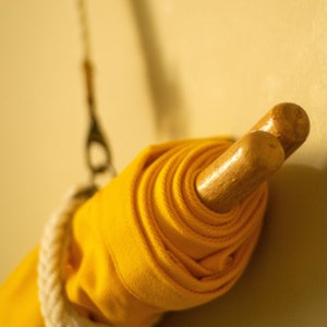 Op de afbeelding: Een gele stof die opgerold en vastgebonden is met een wit touw, hangend aan een houten stok.