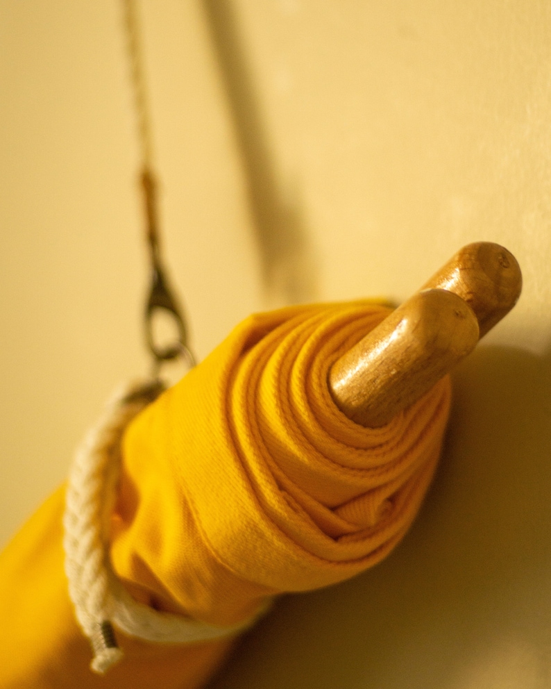 Op de afbeelding: Een gele stof die opgerold en vastgebonden is met een wit touw, hangend aan een houten stok.