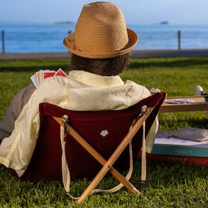 May include: A person wearing a straw hat sits in a red folding chair with a wooden frame. The chair is on a grassy lawn with a body of water in the background.