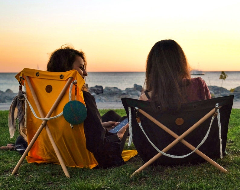Op de afbeelding: Twee mensen zitten op gele en zwarte canvas stoelen met houten frames. De stoelen staan op een grasveld met een waterpartij op de achtergrond. De lucht is licht oranje.