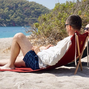Op de afbeelding: Twee mensen ontspannen op een strand in rode en groene opvouwbare stoelen. De stoelen zijn gemaakt van hout en canvas en hebben een eenvoudig ontwerp. De mensen dragen zonnebrillen en casual kleding. Het strand is zanderig en het water is blauw.