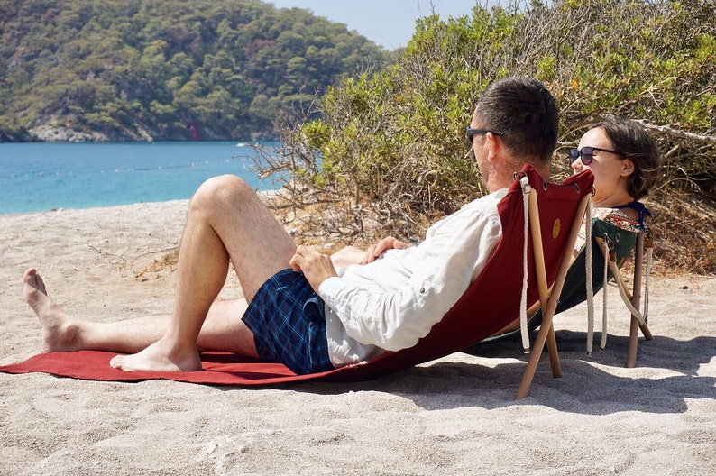 Op de afbeelding: Twee mensen ontspannen op een strand in rode en groene opvouwbare stoelen. De stoelen zijn gemaakt van hout en canvas en hebben een eenvoudig ontwerp. De mensen dragen zonnebrillen en casual kleding. Het strand is zanderig en het water is blauw.