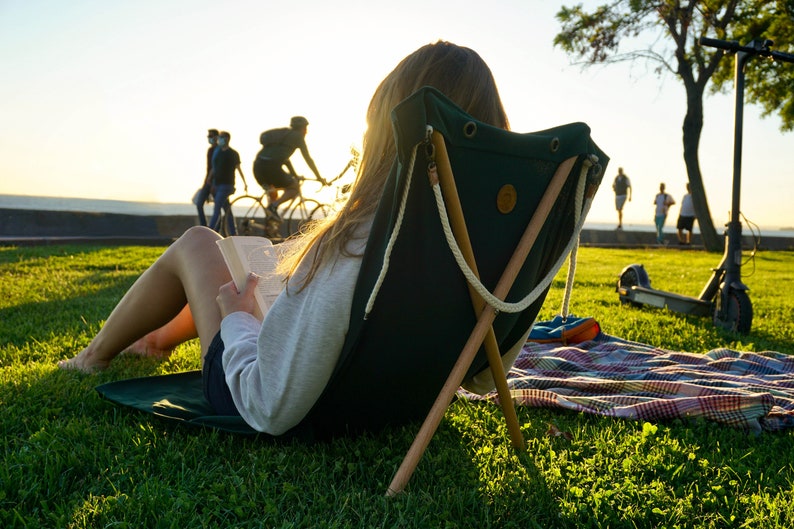 Op de afbeelding: Een persoon zit in een groene canvas vouwstoel met houten poten en leest een boek op een grasveld. De stoel is gemaakt van duurzaam canvas en heeft een eenvoudig ontwerp. De persoon draagt een witte trui en heeft lang bruin haar. Op de achtergrond zijn wazig mensen te zien die fietsen en een scooter rijden.