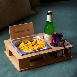 May include: A wooden snack tray with the words "Custom Couchbar" engraved on the side. The tray has a built-in bowl for chips, a bottle holder, and slots for dipping sauces. The tray is sitting on a green surface.