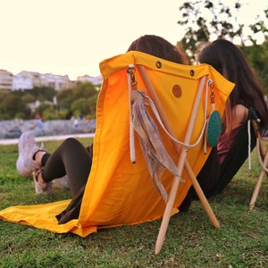 Op de afbeelding: Twee mensen die op gele canvas stoelen met houten poten zitten. De stoelen hebben touwaccenten en een decoratieve kwast. De stoelen staan op een grasveld.
