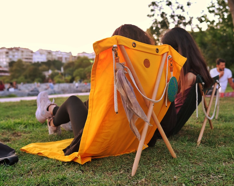Op de afbeelding: Twee mensen die op gele canvas stoelen met houten poten zitten. De stoelen hebben touwaccenten en een decoratieve kwast. De stoelen staan op een grasveld.