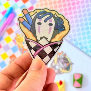 May include: A clear sticker with a cartoon illustration of a No Face character from the movie Spirited Away. The character is inside a crepe with blueberries and chopsticks. The crepe is wrapped in a black and white checkered pattern with a pink stripe.