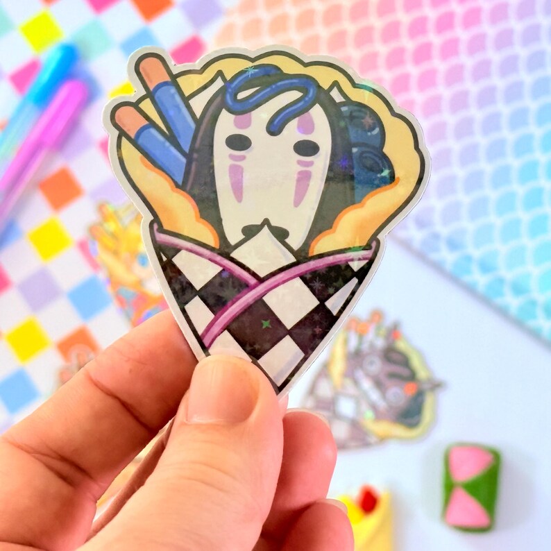 May include: A clear sticker with a cartoon illustration of a No Face character from the movie Spirited Away. The character is inside a crepe with blueberries and chopsticks. The crepe is wrapped in a black and white checkered pattern with a pink stripe.