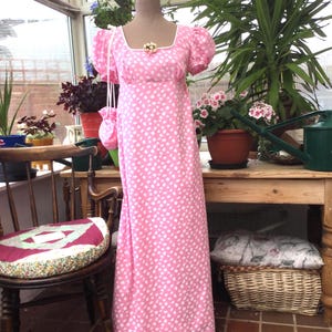 May include: A floor-length pink dress with a white polka dot pattern, featuring short puff sleeves, a square neckline with white trim, and a small floral embellishment at the center chest. A matching small drawstring pouch hangs from the sleeve.