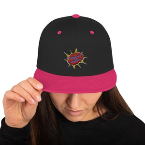 Puede incluir: Gorra de béisbol negra y rosa con una burbuja de diálogo bordada de estilo cómic amarillo y rojo que dice "Cuidado con los explosivos".
