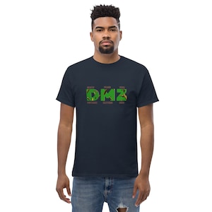Puede incluir: Una camiseta azul marino con un gráfico verde de las letras "DMZ" con las palabras "Death", "Means", "You're", "Distance", "Matters", "Zero" escritas arriba y abajo de las letras.