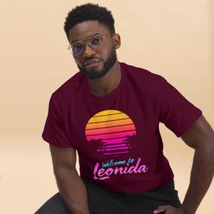 May include: A maroon t-shirt with a retro sunset design. The design includes palm trees and the text "Welcome to Leonida" in a gradient font. The sunset transitions from yellow to orange and pink.