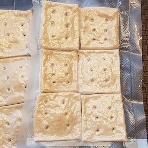May include: Nine square crackers with evenly spaced holes are arranged in a grid, sealed in a clear plastic bag. The crackers are a light tan color, and the bag is partially transparent, revealing the crackers' texture.