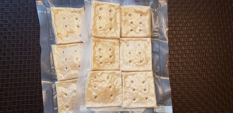 May include: Nine square crackers with evenly spaced holes are arranged in a grid, sealed in a clear plastic bag. The crackers are a light tan color, and the bag is partially transparent, revealing the crackers' texture.