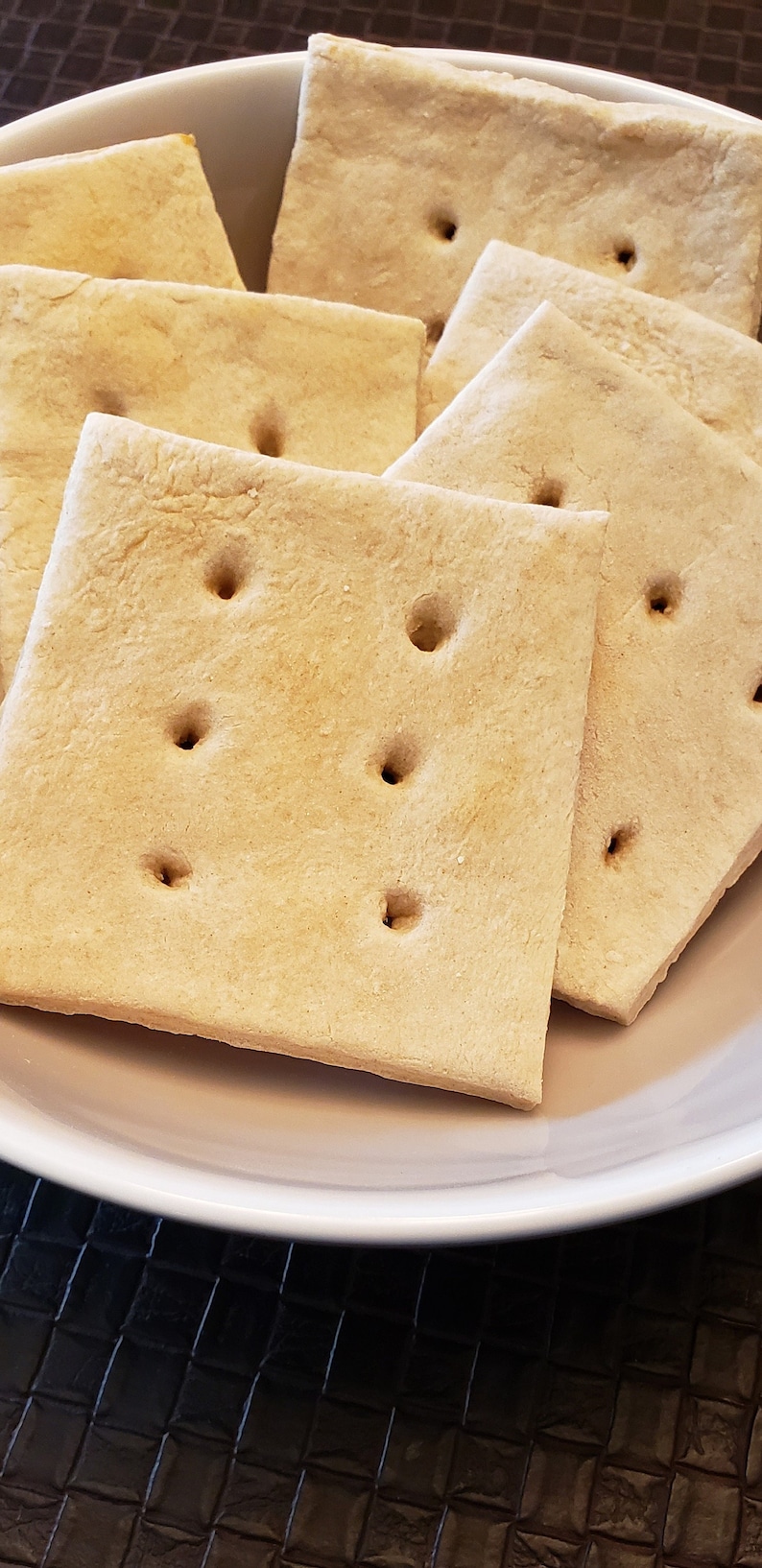 May include: Close-up of square, light brown crackers with four holes each, arranged in a white bowl. The crackers have a slightly rough texture and are stacked on top of each other. The bowl sits on a dark surface.