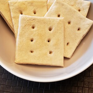May include: A white bowl filled with several square, golden-brown crackers. Each cracker has four small holes. The crackers are arranged in a slightly overlapping pattern, suggesting a snack or appetizer.