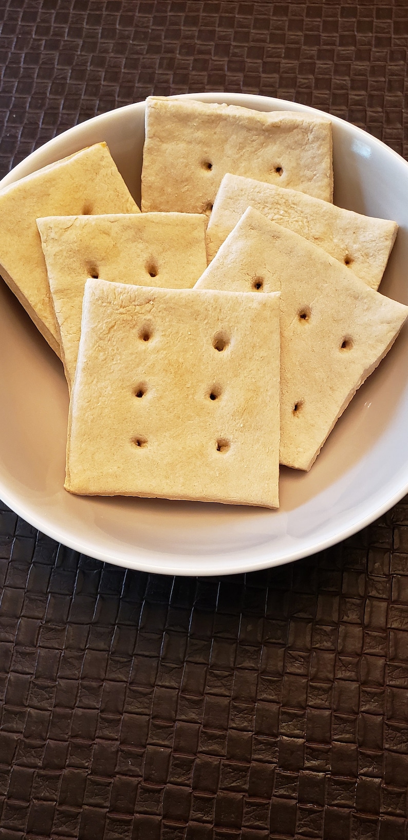 May include: A white bowl filled with several square, golden-brown crackers. Each cracker has four small holes. The crackers are arranged in a slightly overlapping pattern, suggesting a snack or appetizer.
