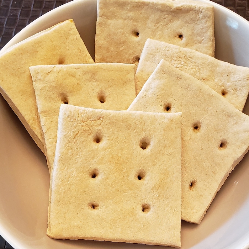 May include: A close-up shot of several square, light brown crackers with evenly spaced holes, arranged in a white bowl. The crackers have a slightly rough texture and appear to be freshly baked. The image highlights the simple, rustic appeal of the snack.
