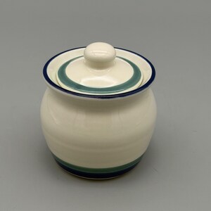 May include: A cream-colored ceramic sugar bowl with a matching lid. The bowl features a dark blue rim and a horizontal band of teal and dark blue stripes near the base. The lid has a small knob handle and a teal circular accent band.