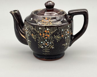 May include: A dark brown ceramic teapot with a matching lid and handle. The teapot is decorated with gold, orange, and white floral patterns. The spout has a gold accent.