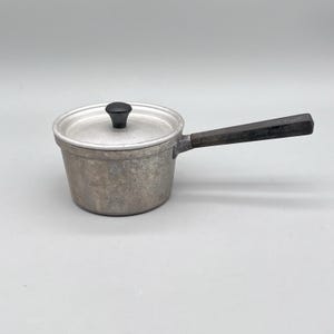 May include: Small, vintage aluminum saucepan with a lid and a long, dark wooden handle. The lid has a black knob. The pot is silver and shows signs of wear, suggesting it's been used. A classic kitchenware item.