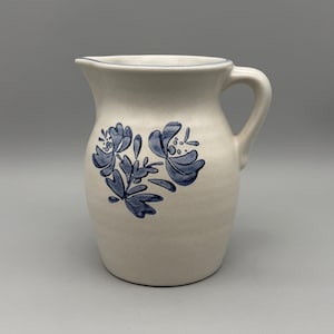 May include: Cream-colored ceramic pitcher with a blue floral design. The pitcher has a handle and a spout for pouring. The blue floral pattern is on the front of the pitcher. The rim of the pitcher is trimmed with a thin blue line.
