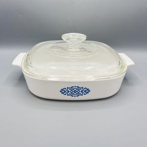 May include: White casserole dish with a clear glass lid and a blue decorative design. The dish has a square shape with rounded edges and two white handles. The lid has a clear knob handle.